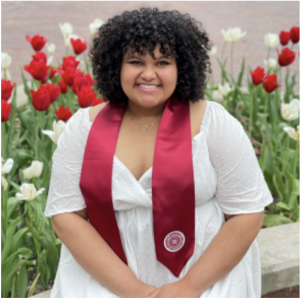 Jenessa Carlisle is pictured wearing a white dress and a red Indiana University graduation stole in front of a patch of tulips. Jenessa is smiling and looking into the camera.